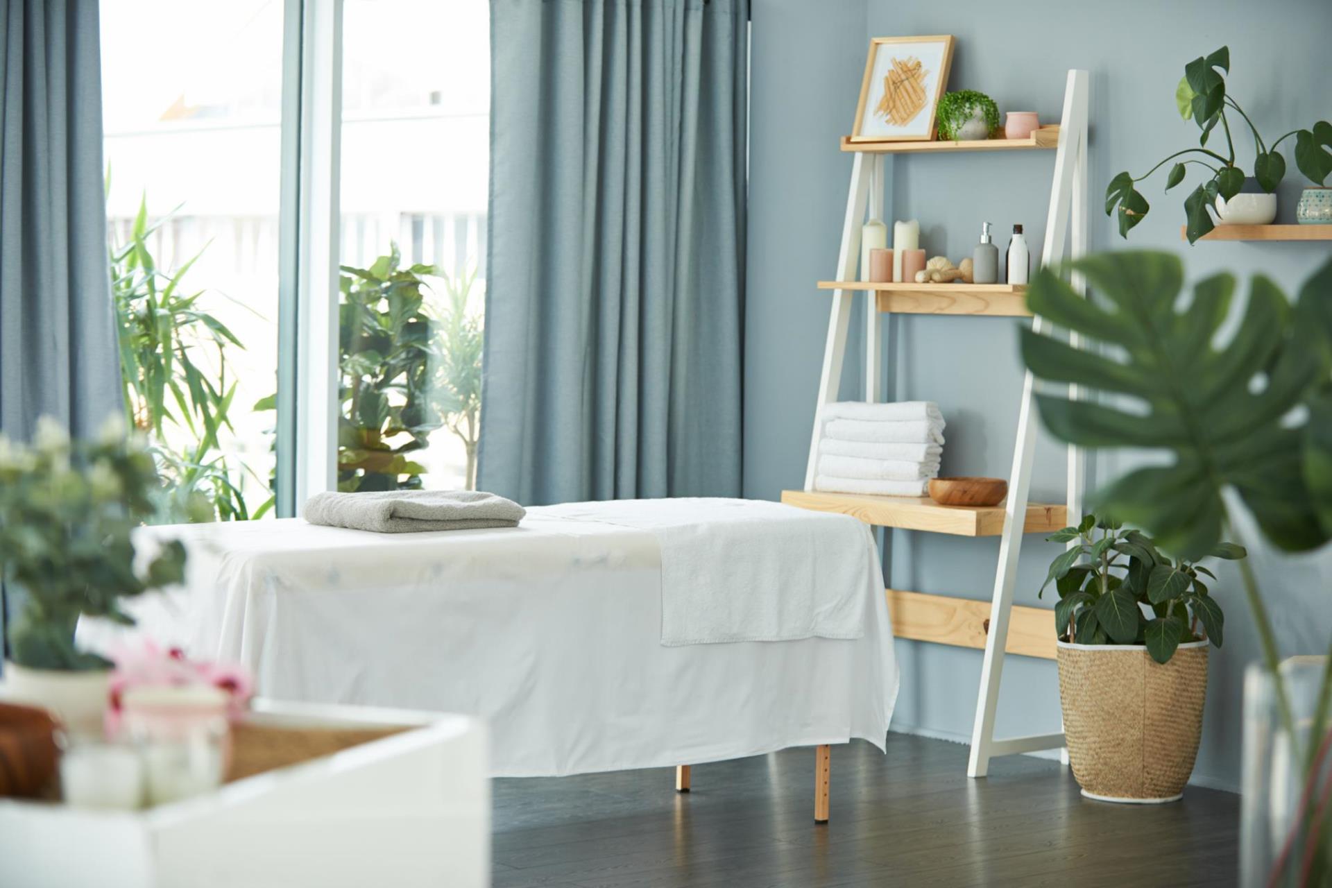 A massage table with table cloth, surrounded by house plants with a shelf nearby